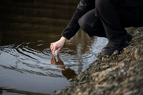 A human hand is holding a sampling bottle, taking a sample from a watercourse.